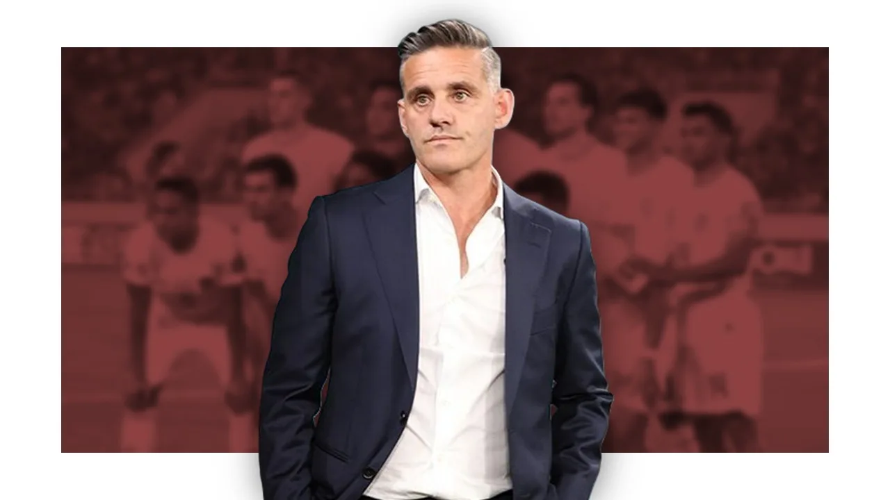 Pelatih sepakbola mengenakan kemeja putih dan jas hitam, menghadap ke kiri, dengan latar belakang foto timnas indonesia dengan efek buram